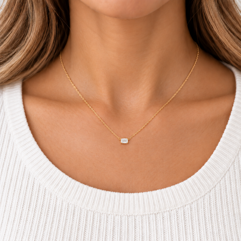 A close-up of a woman's neck and upper chest wearing a delicate gold chain necklace with a small rectangular clear gemstone pendant, paired with a white ribbed knit top.